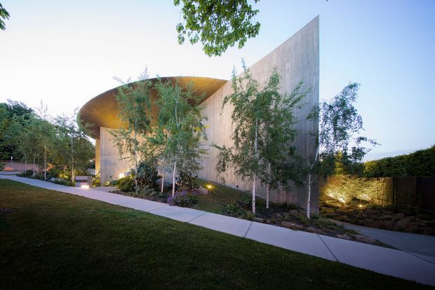 A sculptural modern house exterior set within a landscaped garden. The composition features a tall, sharply angled concrete or rendered wall, a sweeping curved roof or canopy, extensive vertical surfaces, mature planting, illuminated garden beds, and a pale concrete pedestrian path across the foreground. The low evening light highlights the building's geometric form and warm-toned underside.