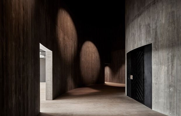 A dramatic, minimalist interior passage or entry space with dark vertical timber wall panelling, a polished concrete floor, and a contrasting raw concrete wall containing a black geometric door. Curved pools of warm light create a moody, sculptural atmosphere, while an opening on the left reveals a brighter adjacent space.