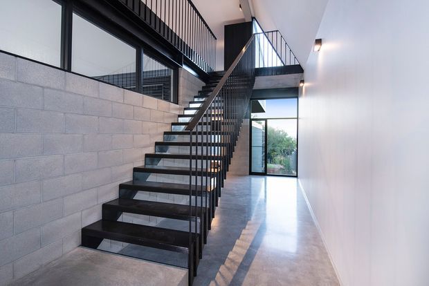 A bright, double-height industrial-style stairway and landing with an open black metal staircase and balustrade, polished concrete flooring, exposed grey concrete block wall, white plaster walls, and large dark-framed glazing. The stair rises to a mezzanine or upper landing, with daylight entering through full-height windows and a glazed door.