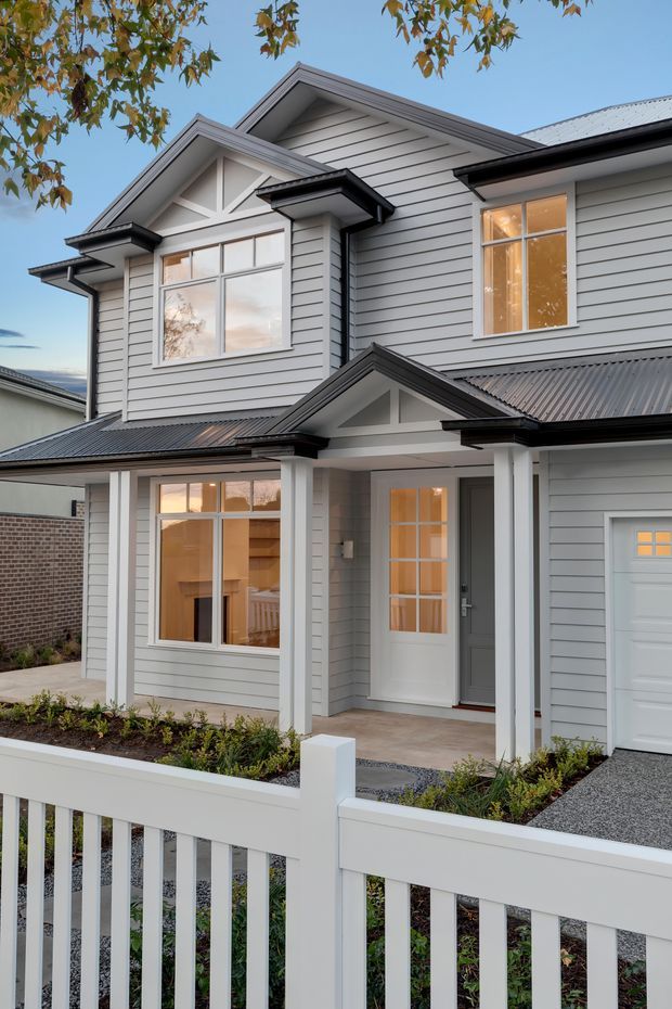 Exterior view of a two-storey suburban house with light grey horizontal weatherboard cladding, dark corrugated metal roofing, white-trimmed multi-pane windows and doors, and prominent gabled roof forms. A covered front porch with white square columns sits beside an integrated garage. The front garden has simple planting, gravel paving, and a white picket-style fence.