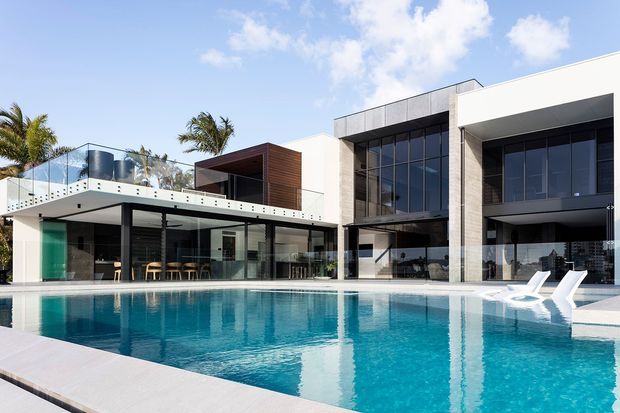 Bright exterior view of a large contemporary multi-level residence with a flat roof, expansive floor-to-ceiling glazing, white rendered walls, dark metal-framed windows, timber-clad feature volumes, and glass balustrades. A broad swimming pool and pool terrace dominate the foreground, with covered outdoor living and dining areas visible beneath the projecting upper levels.