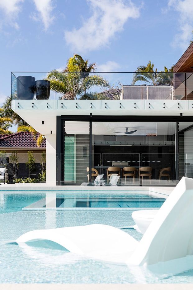 Bright exterior view of a contemporary tropical residence centred on a swimming pool. The house has extensive floor-to-ceiling glazing, a projecting upper terrace with clear glass balustrade and frosted privacy panels, white rendered structural elements, dark-framed openings, and warm timber cladding at the upper right. Palm trees and a tiled roof of a neighbouring or adjacent structure are visible in the background. The pool includes integrated shallow lounging ledges and sculptural white seating or water features.