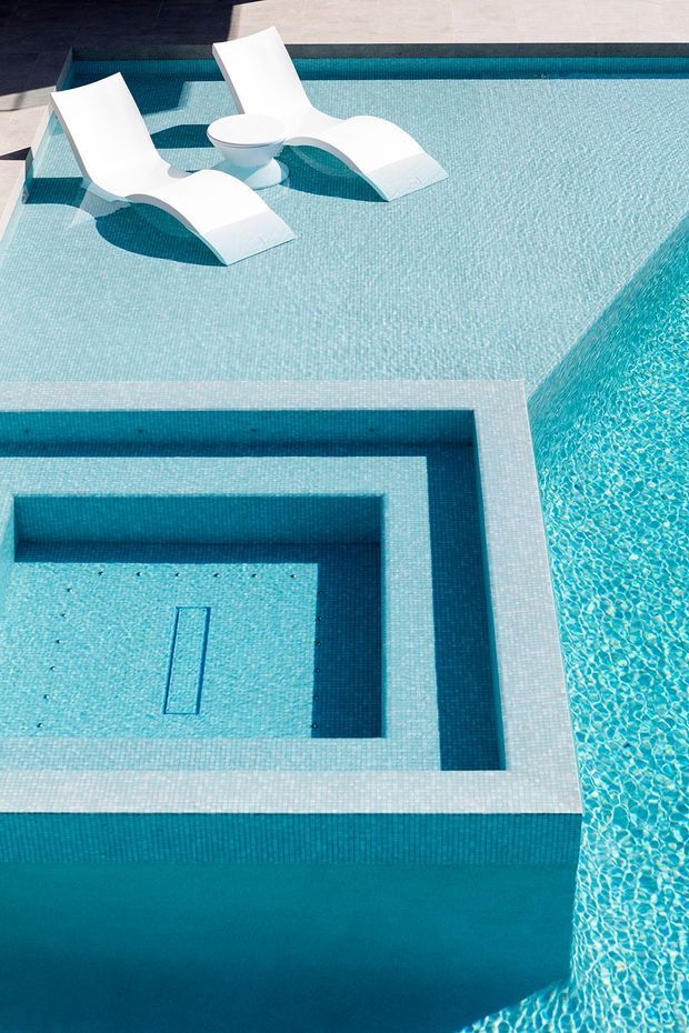 A close-up architectural photograph of a geometric swimming pool with multiple rectangular levels, submerged steps, pale blue mosaic-like tile surfaces, and a raised square spa or shallow inset. Two sculptural white loungers and a small white side table sit on the shallow pool shelf. The image is bright, clean, and strongly focused on the pool design and water geometry.