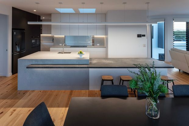 A contemporary open-plan kitchen and dining area with a large grey island, integrated cabinetry, dark appliances, timber flooring, and a linear pendant light. The kitchen features pale grey handleless cabinets, a reflective splashback, stone-look benchtops, skylights, and adjoining living space visible at right. A dark dining table, upholstered chairs, stools, and indoor greenery occupy the foreground.