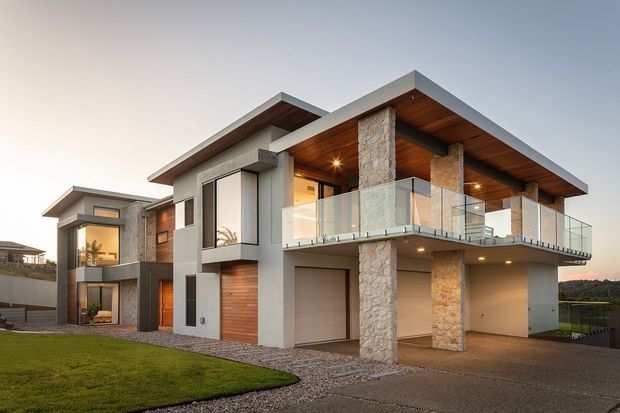 A contemporary multi-level house exterior at dusk, featuring projecting flat roof planes, stone-clad structural columns, smooth rendered walls, timber soffits and wall accents, extensive glazing, and a glass-balustraded upper balcony. A covered carport/garage area and landscaped lawn are visible in the foreground.