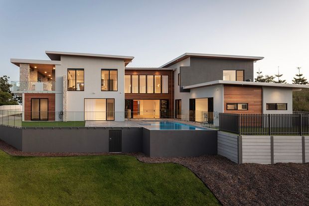 A contemporary multi-level residential house photographed from the landscaped front yard. The façade combines white rendered walls, dark grey rendered volumes, extensive timber-framed glazing, projecting flat rooflines and a central glazed connection. A swimming pool sits within the elevated rear terrace, with dark retaining walls, fencing and landscaped grounds in the foreground.