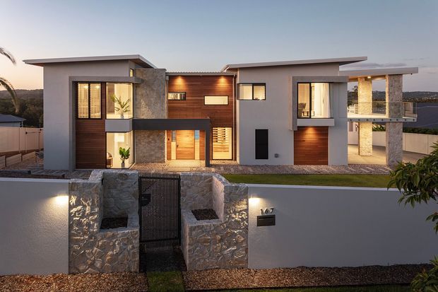 Twilight exterior view of a contemporary multi-level residence with strong horizontal roof planes, white rendered walls, dark timber cladding, and rough natural-stone feature walls and columns. The home has extensive glazing, recessed balconies, a landscaped front boundary, and warm architectural lighting.