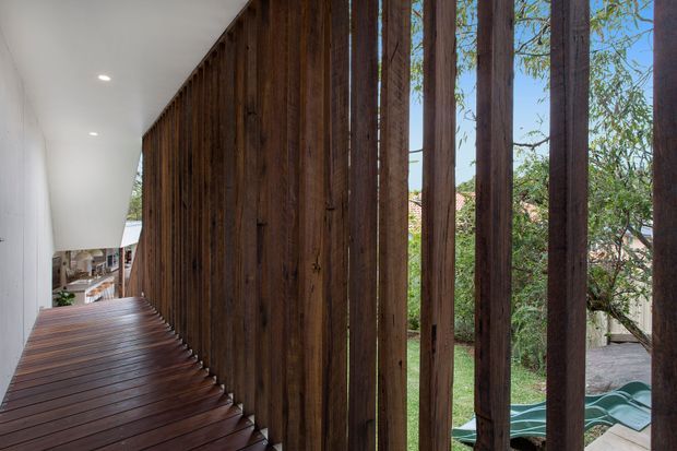 A narrow, modern interior walkway or stair landing with dark timber decking and a dramatic vertical timber screen along one side. A smooth white ceiling with recessed downlights contrasts with the warm wood, while garden greenery and neighbouring buildings are visible through the open slats. The space has a refined indoor-outdoor connection and strong linear architectural detailing.