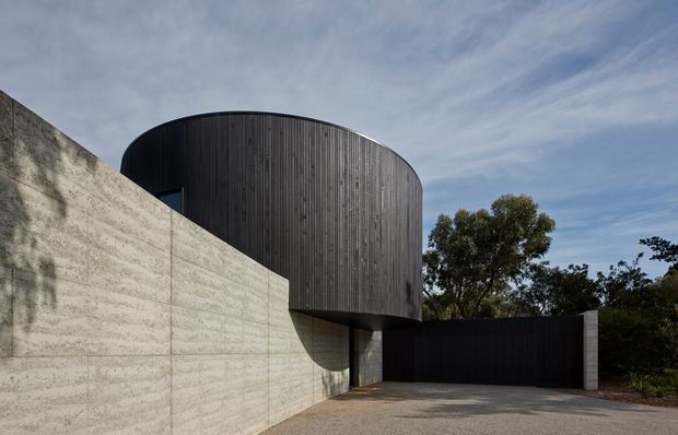 Exterior architectural photograph of a contemporary building with a dramatic rounded upper volume clad in dark vertical timber battens, projecting over a textured pale concrete wall and recessed entrance or garage area. The composition emphasizes curved form, strong horizontal and vertical lines, and restrained dark-and-light material contrast, set against mature trees and a cloudy sky.