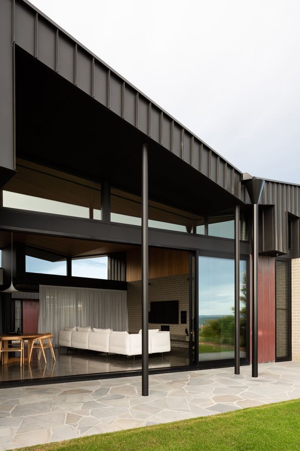 A contemporary residential facade viewed from a paved outdoor terrace, with a deep dark metal roof canopy supported by slender round columns. Full-height black-framed glazing opens onto an interior living room featuring a white sectional sofa, timber dining furniture, warm timber ceiling lining, exposed brickwork, and a fireplace. Irregular pale stone paving, manicured lawn, and a distant landscape are visible outside.