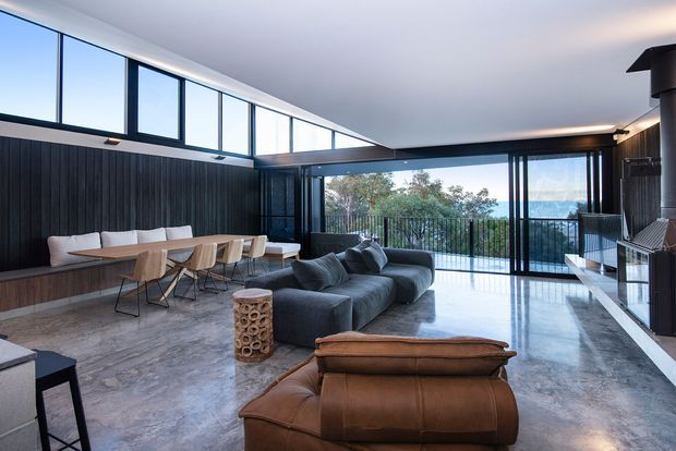 Spacious contemporary open-plan living and dining room with a polished grey stone floor, dark vertical timber feature wall, high clerestory windows, and expansive glazed doors opening to a balcony with a distant water view. A long dining table with built-in bench seating shares the room with a large charcoal sectional sofa, leather lounge chair, and a sleek dark fireplace or media wall. The space has strong natural light, clean linear detailing, and a refined indoor-outdoor connection.