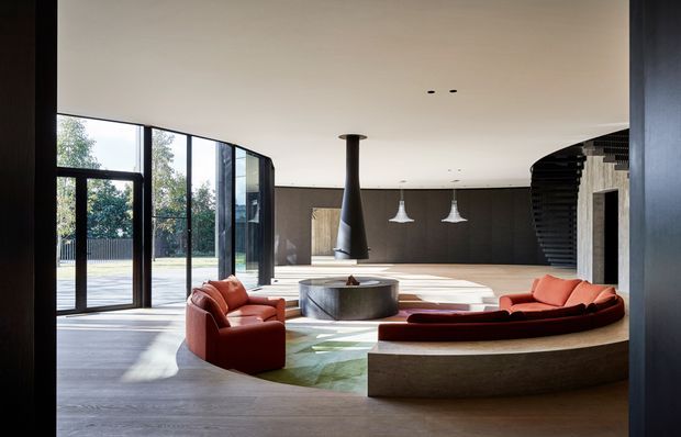 A spacious, high-end contemporary living room with full-height black-framed glazing and a glass door opening to the outdoors. The room features a large curved built-in stone or concrete seating platform with a deep red curved sofa, matching red lounge chairs, a sculptural suspended black fireplace, a round dark coffee table, pale timber flooring, and a dark wood feature wall. A dramatic curved black stair or screening element rises at the right, while white pendant lights hang over a partially visible kitchen or dining zone.