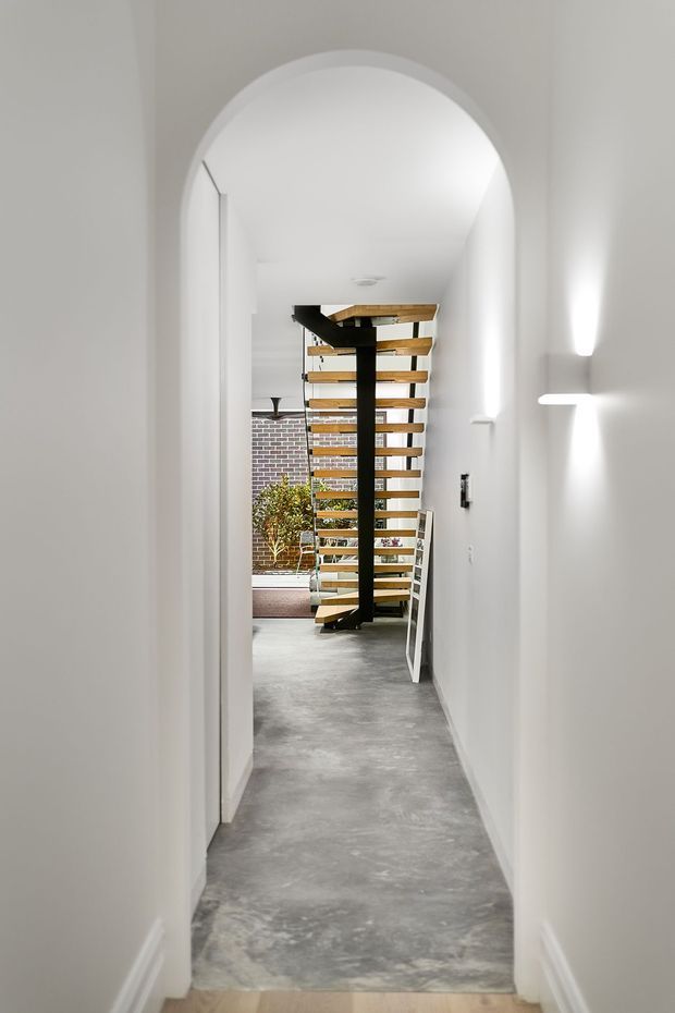 A narrow, bright hallway viewed through a tall rounded archway, leading to an open-riser timber staircase with a prominent black central steel support. White walls and ceiling contrast with a grey polished concrete floor, while exposed brick and indoor planting are visible beyond the stairs. Minimal wall lighting and a clean, contemporary palette create a refined transitional circulation space.