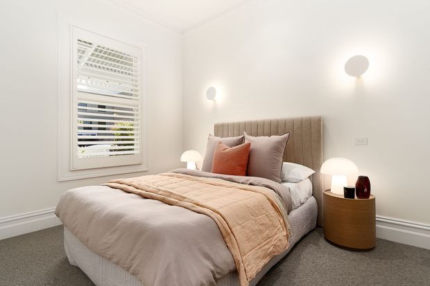A bright, softly styled bedroom with a double bed, upholstered vertical-panel headboard, layered neutral bedding, and muted terracotta and taupe cushions. The room has pale walls, grey carpet, white plantation shutters, wall-mounted sconces, and a rounded timber bedside table with a table lamp and decorative vessel.