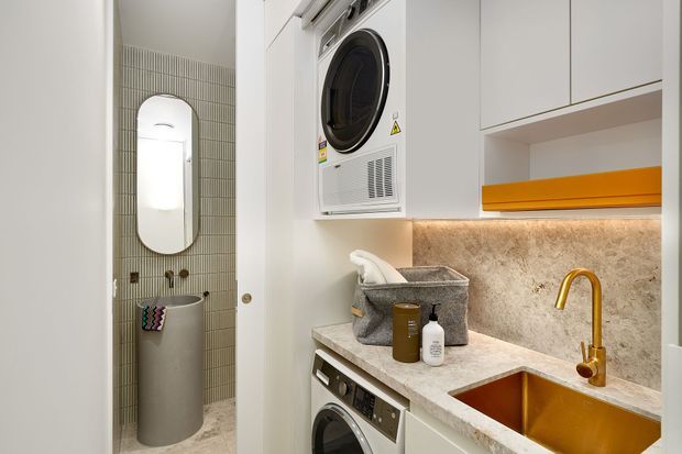 Compact contemporary laundry with stacked front-loading washer and dryer, white overhead cabinetry, a stone-look benchtop and splashback, and a gold-toned sink and tap. A tiled utility area with a freestanding hamper and wall-mounted mirror is visible through the opening on the left.