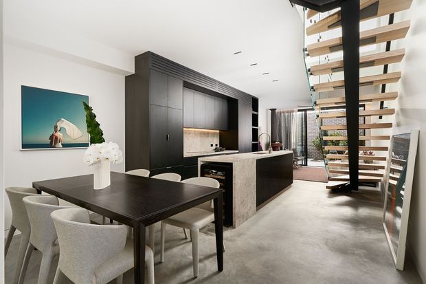 Open-plan contemporary kitchen and dining area with a dark timber dining table, upholstered dining chairs, large black built-in cabinetry, a stone island bench, and a floating timber staircase with black supports. The bright interior has polished concrete flooring, white walls, and restrained modern furnishings.