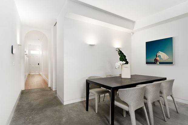 A bright, minimalist dining area with a dark rectangular dining table and upholstered chairs, set against white walls with concealed or subtle wall lighting. The room opens to a hallway with an arched doorway and white entry door. Grey polished concrete flooring, simple ceiling beams, a potted plant, and a framed artwork complete the restrained contemporary interior.