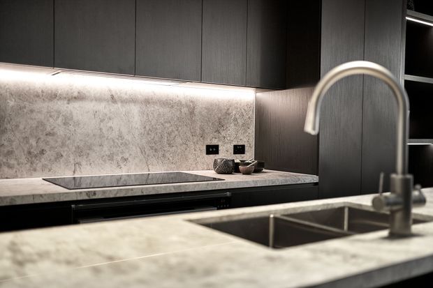 A contemporary kitchen viewed across a large stone island benchtop, featuring a deep undermount sink and tall brushed metal gooseneck tap. Dark vertical-grain cabinetry runs along the back wall, with integrated appliances, a pale veined stone splashback and benchtops, under-cabinet lighting, and open shelving at right. The overall scheme is dark, refined, and highly streamlined.