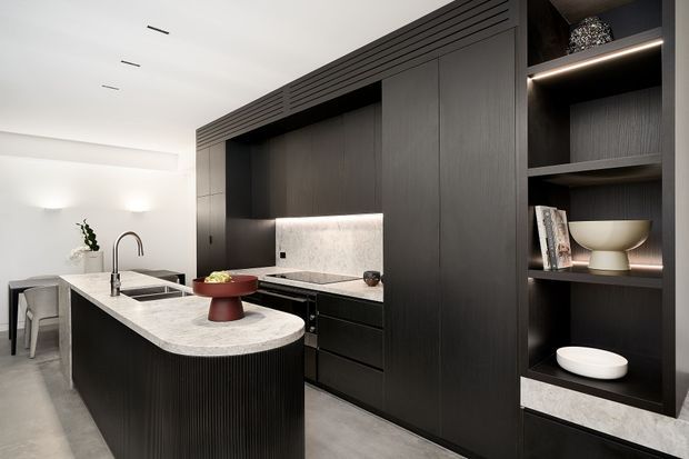 A contemporary kitchen with a large rounded island, dark vertically fluted cabinetry, integrated black appliances, and a pale stone benchtop. A softly illuminated stone splashback sits behind the cooktop, while open display shelving with concealed lighting adds a refined decorative element. The space has light grey flooring, white walls, minimalist lighting, and a clean, monochromatic palette.