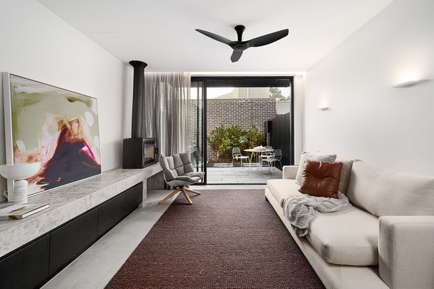 Contemporary living room with a large cream sectional sofa, dark textured rug, black built-in media cabinetry and a pale stone television console. A sculptural black ceiling fan, wall-mounted lights and a suspended black fireplace flue add modern detailing. Full-height sliding glazing opens to a small paved courtyard with brick screening and outdoor furniture.