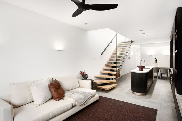 Bright open-plan contemporary interior combining a living room, kitchen and dining area. A floating timber staircase with glass balustrade leads upward beside a compact kitchen island, while white walls, polished grey flooring, a large cream sofa and dark rug create a clean, minimalist feel. A black ceiling fan and dark cabinetry provide contrast.
