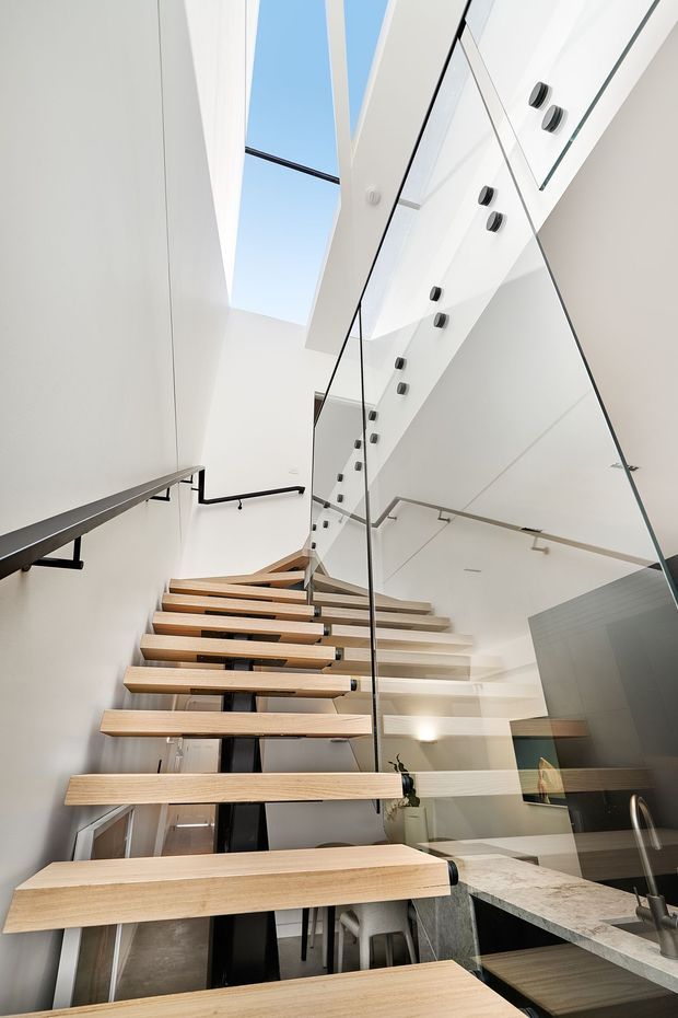 A bright, narrow contemporary interior featuring an open-riser timber staircase with floating wooden treads, black metal supports and handrails, and clear glass balustrades. The stair rises beside a compact kitchen with pale stone benchtops and dark cabinetry. White walls and ceilings, extensive glazing or skylight-like openings, and mirrored or reflective surfaces create a tall, airy feel.