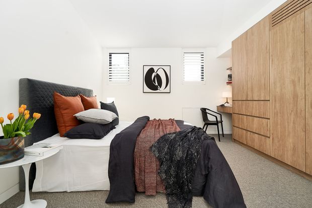 Bright, contemporary bedroom with a neatly made bed, dark upholstered headboard, rust and charcoal bedding, white walls, plantation shutters, abstract artwork, and extensive light timber built-in cabinetry along one wall. A small desk and chair sit beside the cabinetry, with a side table and flowers near the bed.