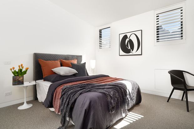 Bright, compact contemporary bedroom with a dark upholstered quilted headboard, layered charcoal and rust bedding, white walls, carpeted flooring, two louvered windows, a small round side table with flowers, and a minimalist desk chair. Abstract black-and-white artwork adds a graphic accent.