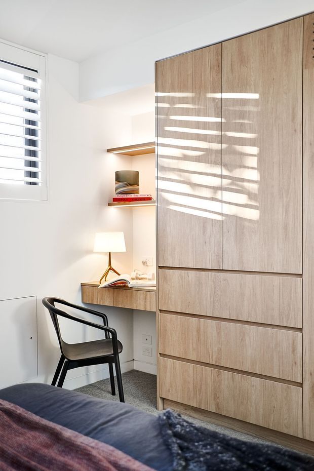 Bright compact bedroom with a built-in timber wardrobe occupying the right side, a small integrated study desk and open shelves, a black armchair, bedside lamp, and part of the bed in the foreground. White walls, a plantation-shutter window, and sunlight filtering through the slats create a clean, warm, contemporary feel.