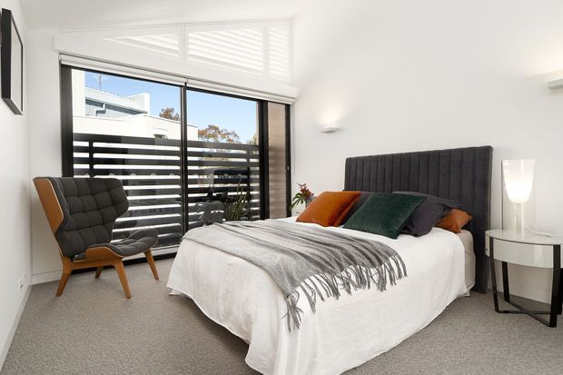 Bright contemporary bedroom with a large upholstered bed, neutral bedding, colourful accent cushions, a sculptural lounge chair, and a compact round bedside table. Full-height sliding glass doors open to a balcony with horizontal screening, while white walls, carpet flooring, and streamlined lighting create an airy, refined interior.