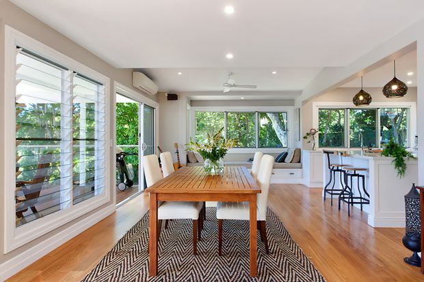 Bright open-plan dining and kitchen interior with a timber dining table and upholstered chairs on a patterned rug, opening to a white kitchen with a breakfast bar and pendant lights. Large louvred windows and sliding glass doors bring in greenery, while timber flooring, recessed lighting, a ceiling fan, and built-in window seating create a relaxed tropical-coastal feel.