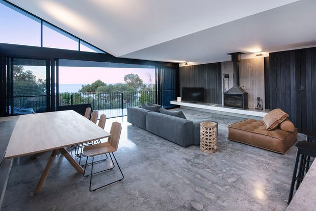 Open-plan contemporary living and dining room with a dramatic angled ceiling, extensive floor-to-ceiling glazing, and access to a balcony or terrace. The room features a large dining table, sectional sofa, television, sculptural fireplace surround, dark vertical wall panelling, and a polished concrete-look floor. The palette is predominantly cool grey, black, and neutral timber tones, with views to greenery beyond the glazing.