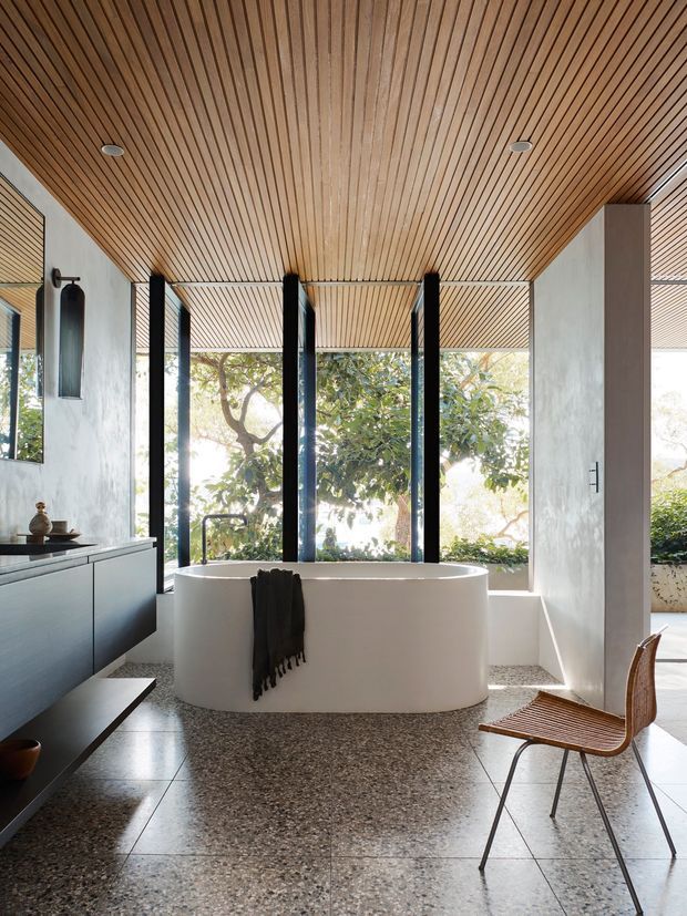 A bright, contemporary bathroom with a freestanding white oval bathtub positioned before full-height glazing. The room features a richly lined timber slat ceiling, pale textured walls, dark-framed windows, a dark floating vanity, and a terrazzo floor. A simple wooden chair and towel add understated warmth, while leafy garden views are visible outside.