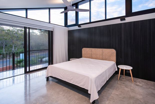 A bright contemporary bedroom with a low platform bed, white bedding, rounded upholstered headboard, and small round bedside tables. The room has polished concrete flooring, dark vertical wall panelling, expansive black-framed glazing, a high glazed ceiling, and a ceiling fan. Outdoor vegetation and a balcony are visible beyond the glass.