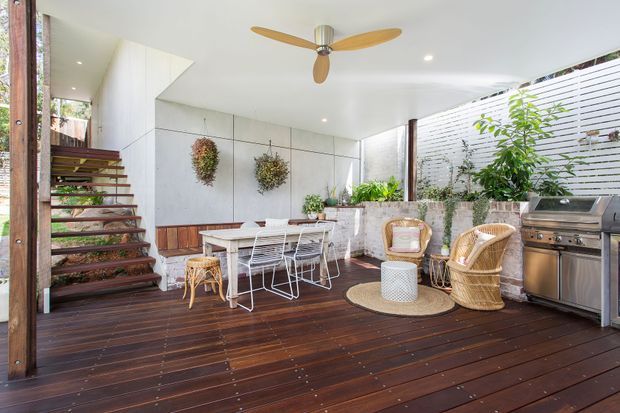 A bright, covered outdoor entertaining area with a dark timber deck, dining table and chairs, wicker lounge seating, built-in planter beds, barbecue, timber posts, and an open stair leading to an upper level. The space has a white ceiling with recessed lights and a timber-bladed ceiling fan, while planted greenery and a rendered masonry privacy wall create a lush courtyard atmosphere.