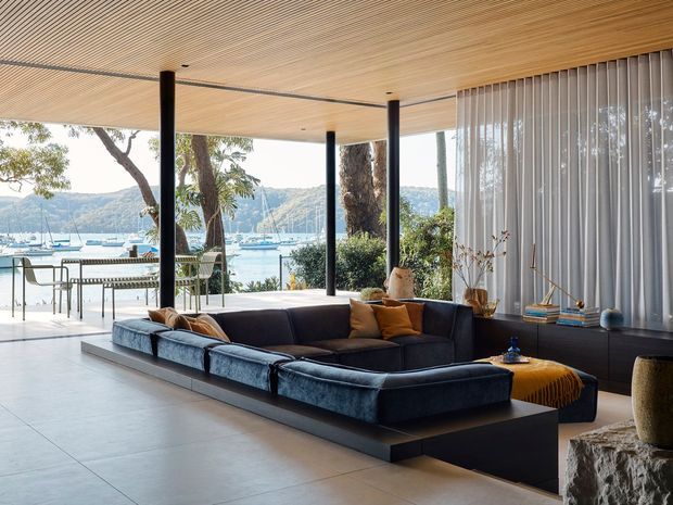 A refined contemporary living room with a large dark blue modular sectional sofa, low built-in cabinetry, and a mustard upholstered ottoman. The room opens through extensive floor-to-ceiling glazing onto a bright waterfront setting with boats and hills visible beyond. A warm timber slatted ceiling, slim black structural columns, pale large-format floor tiles, and sheer vertical blinds create a relaxed indoor-outdoor atmosphere.