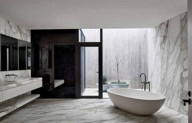 A luxurious contemporary bathroom featuring a freestanding oval bathtub, wall-mounted double vanity, black-framed glazing, and extensive white-and-grey veined marble surfaces. A dark tiled shower area is visible at the rear, with a small planted courtyard beyond the glazing.