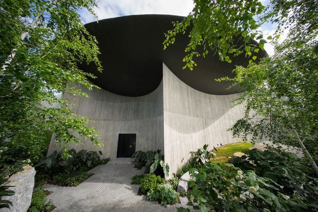 Exterior view of a sculptural, monolithic concrete building set within dense landscaped greenery. Curved board-formed concrete walls rise beneath a dramatic dark, sweeping roof canopy, with a small black entrance door reached by a paved walkway. The composition emphasizes organic curves, strong shadows, and integration with the surrounding garden.