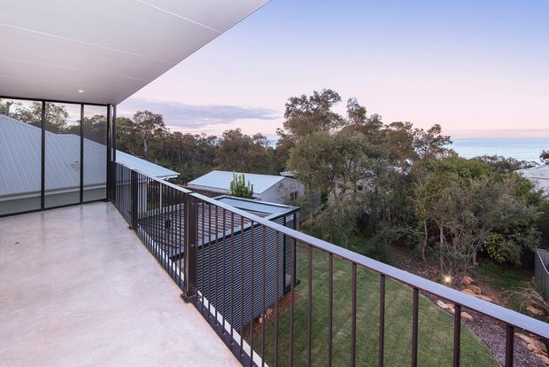 A spacious elevated exterior terrace or balcony with a smooth concrete floor, black metal balustrade, and broad flat roof overhang. The terrace overlooks trees, neighbouring rooftops, and a visible coastal body of water in the distance. The setting has a contemporary architectural character with expansive glazing and a calm dusk atmosphere.
