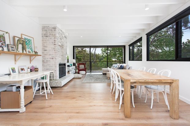 Bright open-plan living and dining room with extensive black-framed glazing, white-painted exposed ceiling beams, pale timber flooring, a whitewashed brick fireplace surround, and a large rustic timber dining table with white chairs. The space includes a lounge area, fireplace, shelving, artwork, and views to trees outside.