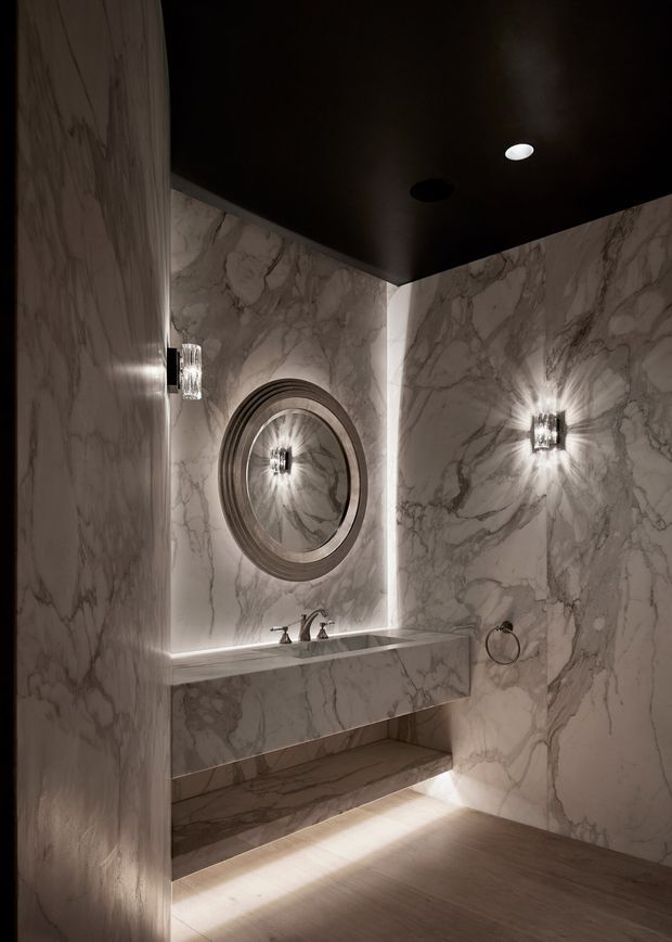 A compact, dramatic bathroom clad almost entirely in veined marble, with a floating marble vanity, integrated open shelf, traditional metal tapware, circular framed mirror, wall sconces, and concealed under-vanity and mirror lighting. The dark ceiling and warm lighting create a luxurious, moody atmosphere.