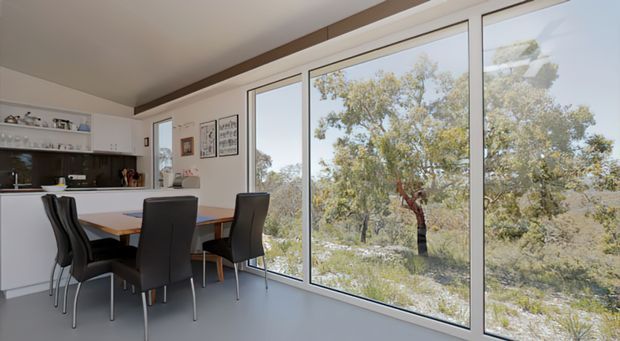 Bright open-plan dining area beside a compact kitchen, with a rectangular timber dining table and dark upholstered chairs set against expansive floor-to-ceiling windows. The room has white cabinetry and walls, a dark stone-look kitchen benchtop, a smooth pale grey floor, and a timber ceiling beam or trim. Large glazing overlooks a natural bush landscape.