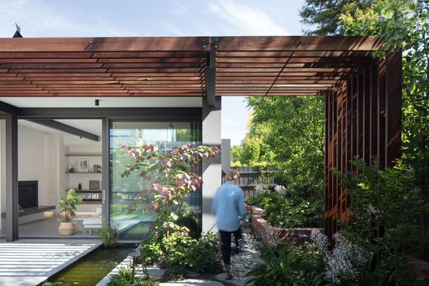A contemporary residential courtyard viewed from outdoors, featuring a dark timber pergola and vertical timber screening framing a glazed living area. Lush planted garden beds, a narrow reflecting or water channel, paved paths, and a partially visible minimalist interior create a layered indoor-outdoor setting.