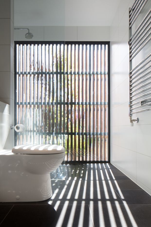 A bright contemporary bathroom with a wall-mounted toilet, white tiled walls, dark floor tiles, and a large glazed opening screened by vertical slats. Strong sunlight creates dramatic striped shadows across the floor and toilet. A chrome heated towel rail is mounted on the right wall, with a minimalist shower fitting visible on the left.