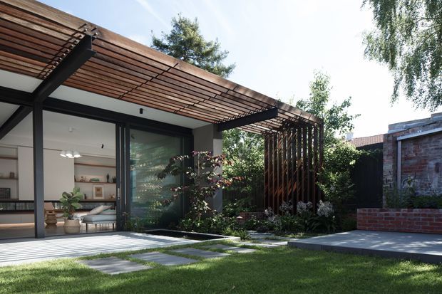 A contemporary residential courtyard viewed from the garden, featuring a dark steel-framed glazed extension beneath a timber slatted pergola. The pergola continues as a vertical timber screen around a planted garden edge. Large sliding glass doors reveal a calm living area inside, while concrete stepping slabs, lawn, raised brickwork, and an exposed brick boundary wall define the outdoor setting.