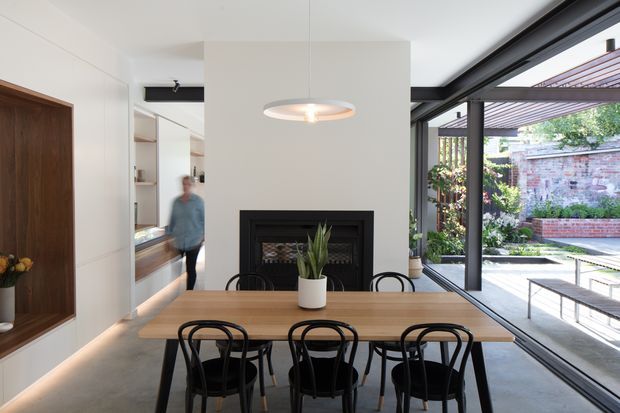 A bright contemporary dining area with a long timber dining table and black bentwood-style chairs, positioned beside a black-framed fireplace and extensive glazed openings. A built-in timber display niche and shelving line the left wall, while the glazing opens to a landscaped brick courtyard with a pergola and outdoor seating.