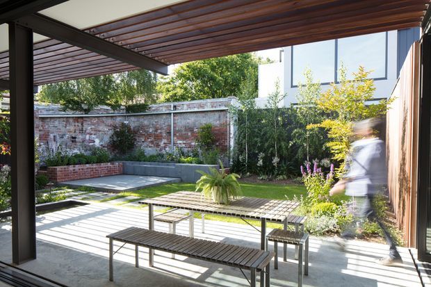 A bright landscaped courtyard or patio viewed from beneath a timber-slat pergola. The space features a concrete-paved entertaining area, simple slatted outdoor tables and benches, raised brick and concrete garden beds, established planting, and an aged brick boundary wall. Dark structural framing and a contemporary building are visible around the courtyard, with strong sunlight casting linear shadows across the paving.