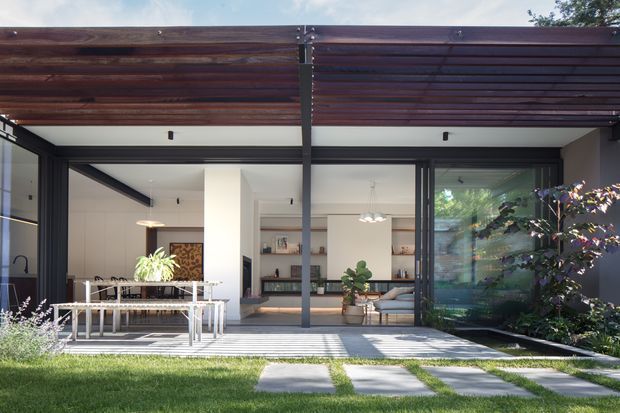 Contemporary residential courtyard view looking toward a house through a covered outdoor terrace. A dark steel pergola with timber slats shades the patio, which contains an outdoor dining table and chairs. Large dark-framed glazed openings reveal a bright open-plan interior with dining and living areas, built-in shelving, plants, and neutral finishes. The foreground includes a lawn and stepping-stone path, with established planting along the right side.