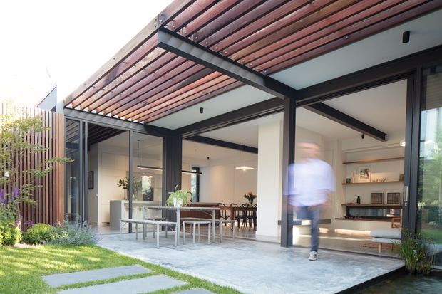 Contemporary single-storey home opening onto a landscaped patio through extensive dark-framed glazing. A timber slatted pergola extends over the outdoor terrace, with an open-plan dining and living area visible inside. The design combines warm timber overhead screening, dark structural framing, pale walls, and a light paved terrace, creating a bright indoor-outdoor connection.