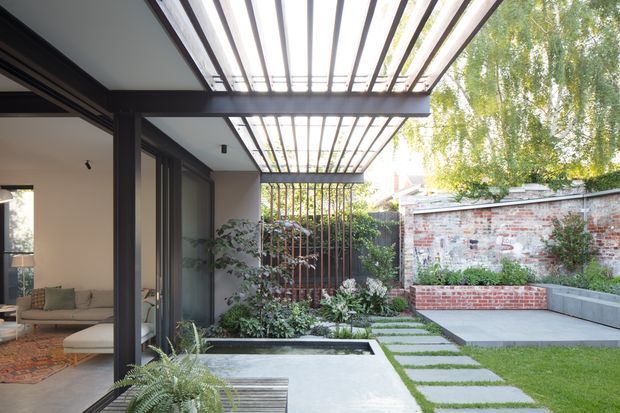 A contemporary courtyard garden viewed from a covered outdoor patio beside a glazed living area. A dark steel pergola with closely spaced slats creates a strong overhead rhythm, while stepping stones cross pale paving and lawn toward raised brick and concrete garden beds. Exposed brick boundary walls, dense planting, and a narrow rectangular water feature frame the calm outdoor space. Part of the adjoining living room with a sofa is visible on the left.